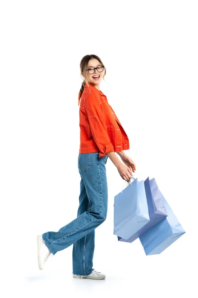 Pleine longueur de femme gaie en chemise orange tenant des sacs à provisions isolés sur blanc — Photo de stock