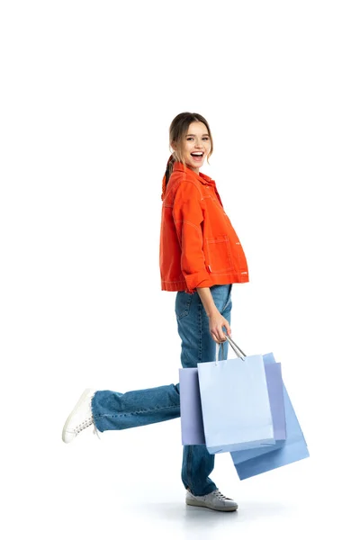 Volle Länge der jungen positiven Frau in orangefarbenem Hemd mit Einkaufstaschen isoliert auf weiß — Stockfoto