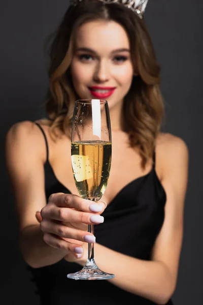 Mulher feliz borrada em vestido de deslizamento preto e tiara segurando copo de champanhe em cinza — Fotografia de Stock