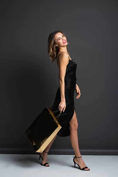 Pleine longueur de jeune femme heureuse en robe noire glissante tenant sac à provisions brillant et marchant sur gris — Photo de stock