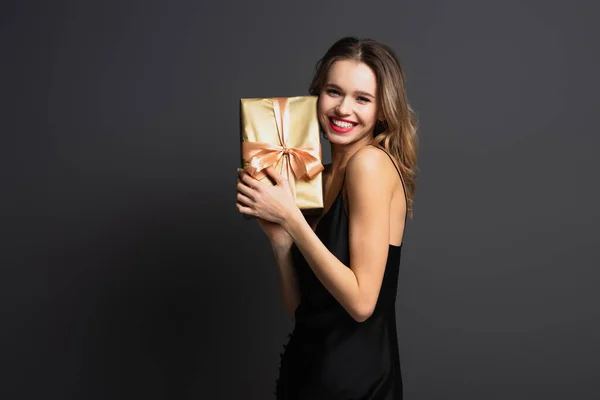 Happy young woman in black slip dress holding wrapped gift box on grey — Stock Photo