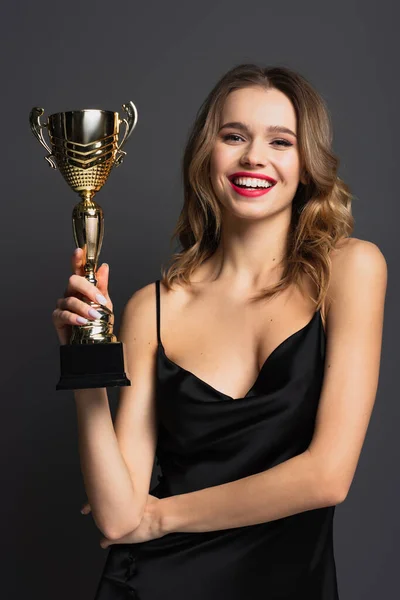 Heureuse jeune femme en robe noire glissante tenant trophée d'or sur gris — Photo de stock