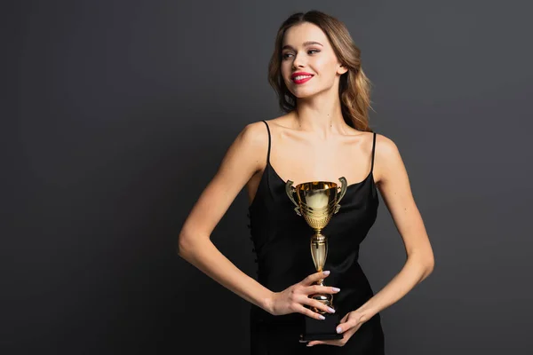 Веселая молодая женщина в черном платье с золотым трофеем на сером — стоковое фото