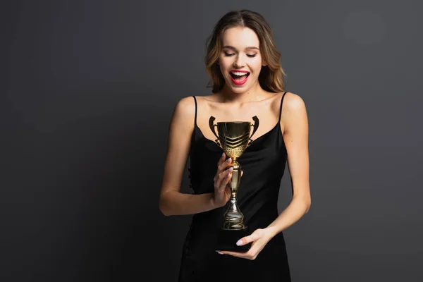 Excitée jeune femme en robe noire glissante tenant trophée d'or sur gris — Photo de stock