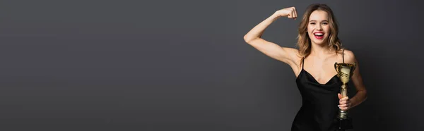 Happy young woman in black slip dress holding golden trophy and showing muscle on grey, banner — Stock Photo