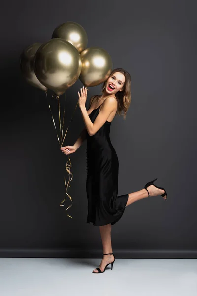 Pleine longueur de jeune femme étonnée en robe noire glissante tenant des ballons dorés sur gris — Photo de stock