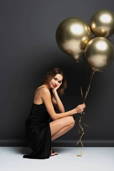 Longitud completa de la joven feliz en vestido negro deslizante sosteniendo globos de oro mientras está sentado en gris - foto de stock