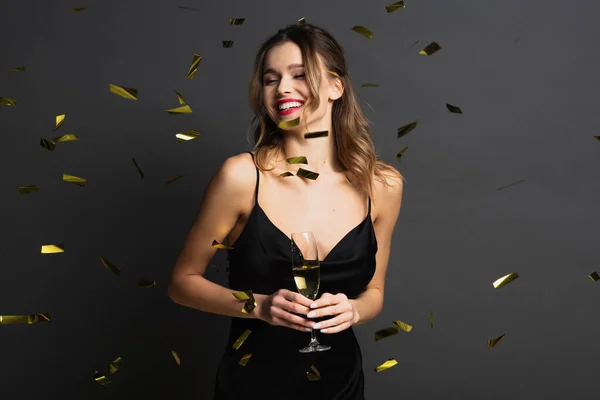 Joyeuse jeune femme en robe noire glissante tenant un verre de champagne près de confettis sur gris — Photo de stock