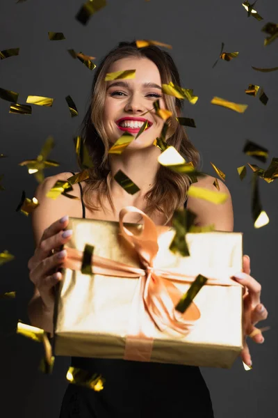 Gioiosa giovane donna in abito antiscivolo nero contenente confezione regalo vicino confetti sfocati sul grigio — Foto stock