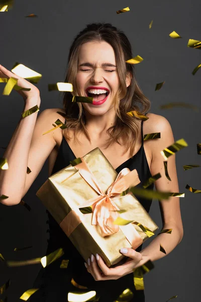 Eccitata giovane donna in abito antiscivolo nero contenente confezione regalo vicino confetti sfocati sul grigio — Foto stock