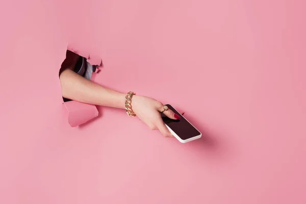 Vista recortada de la mujer con teléfono inteligente en la mano cerca de fondo rosa con agujero - foto de stock