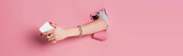 Partial view of young woman holding smartphone in hole of pink background, banner — Stock Photo