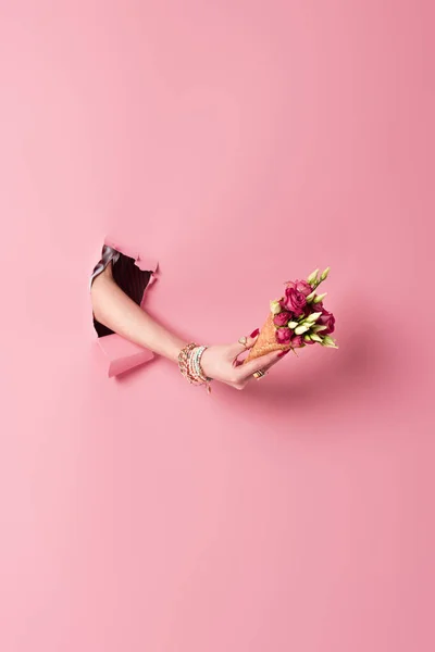 Vista recortada del cono de gofre con flores en la mano de la mujer en el agujero de fondo rosa - foto de stock