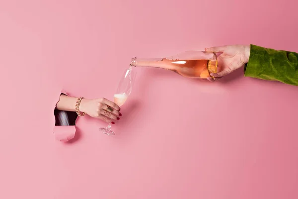 Vista recortada del hombre vertiendo champán cerca de la mujer con vidrio y fondo rosa con agujero - foto de stock