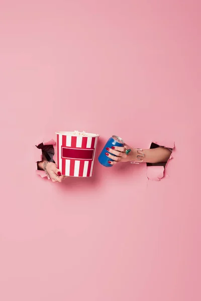 Cropped view of woman holding canned drink and popcorn in holes of pink background — Stock Photo