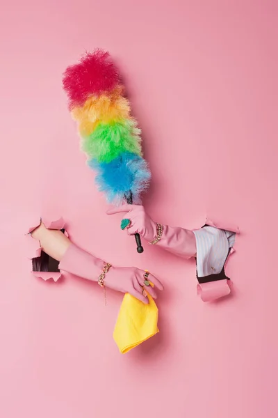 Cropped view of woman in gloves holding rag and dust brush near pink background with holes — Stock Photo