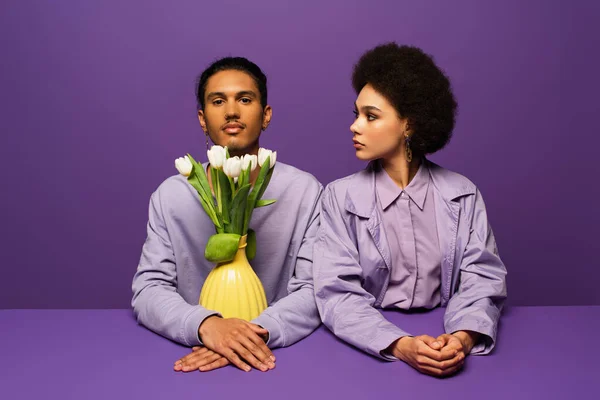 Jeune couple afro-américain assis près du vase avec des tulipes isolées sur violet — Photo de stock