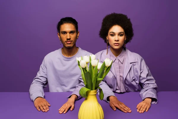 Casal afro-americano sentado perto do vaso com tulipas brancas isoladas em roxo — Fotografia de Stock