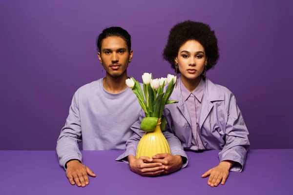 Jovem e moderno casal afro-americano sentado perto do vaso com tulipas isoladas em roxo — Fotografia de Stock