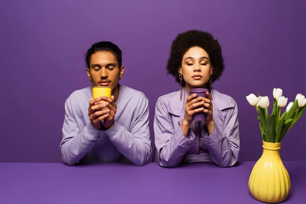 African american couple sitting near vase with white tulips and holding paper cups isolated on purple — Stock Photo