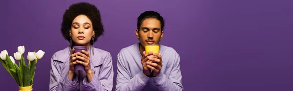 African american couple sitting near vase with white tulips and holding paper cups isolated on purple, banner — Stock Photo