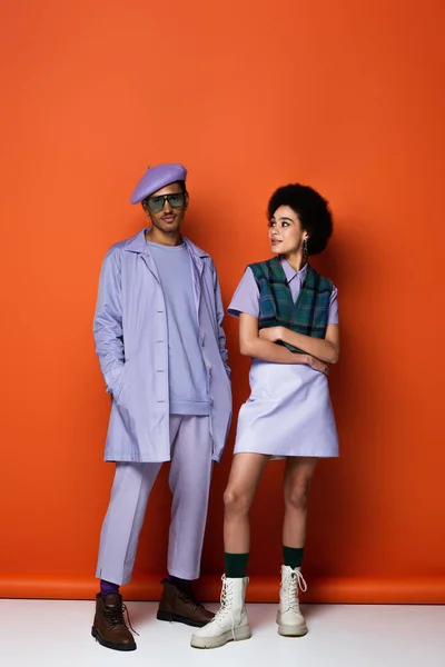 Longitud completa de la mujer afroamericana de moda de pie con los brazos cruzados y mirando a un hombre con estilo en naranja - foto de stock