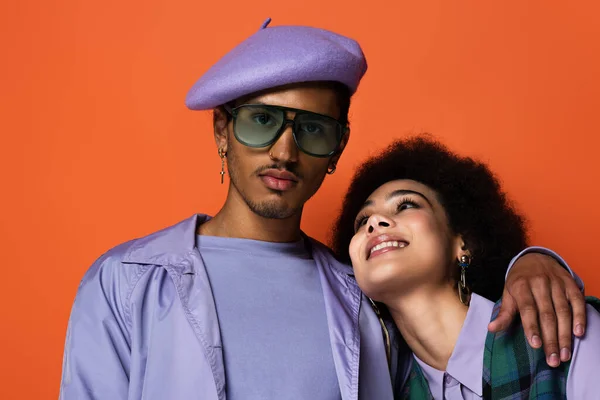 Homme afro-américain à la mode en béret et lunettes de soleil étreignant femme heureuse isolée sur orange — Photo de stock