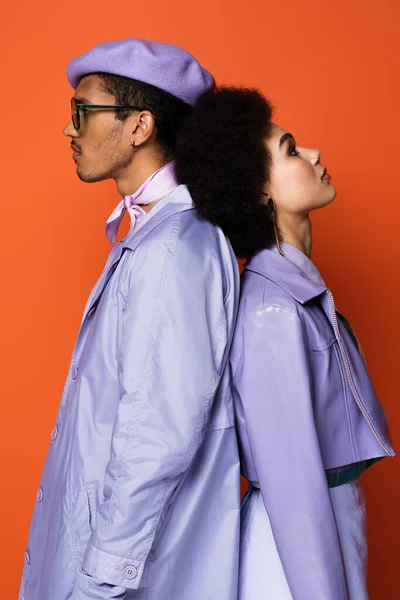 Femme bouclée en veste violette debout dos à dos avec homme élégant en béret isolé sur orange — Photo de stock