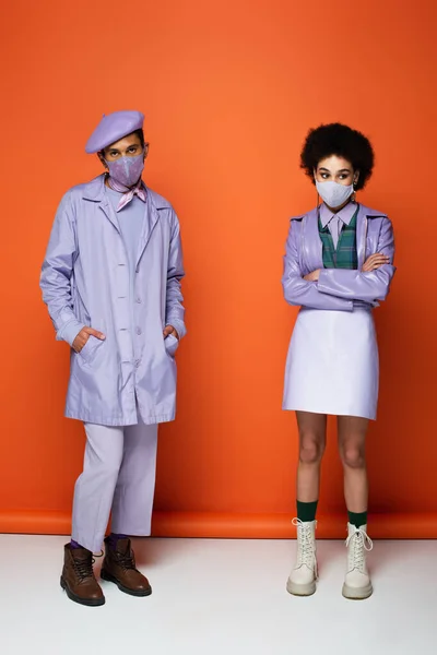 Full length of trendy african american couple in protective masks standing on orange — Stock Photo