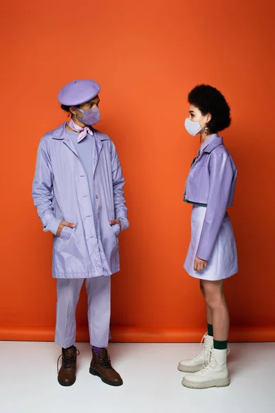 Comprimento total de casal americano africano na moda em máscaras protetoras olhando um para o outro em laranja — Fotografia de Stock