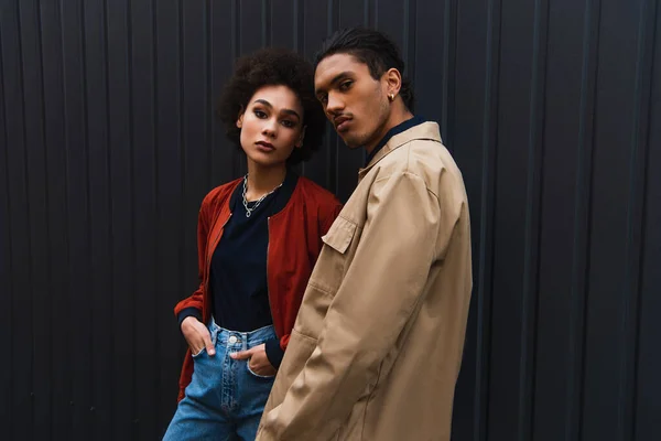 Joven afroamericano hombre mirando la cámara mientras posando con elegante mujer de pie con las manos en los bolsillos - foto de stock