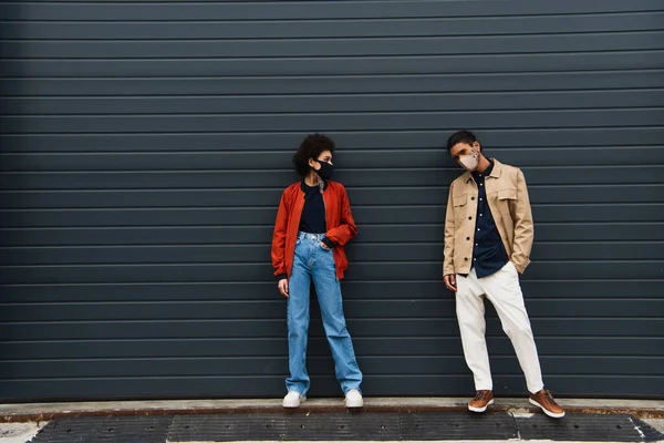 Full length of young african american woman and stylish man in protective masks posing with hands in pockets outside — Stock Photo