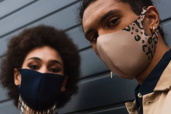 Hombre afroamericano joven y mujer borrosa en elegantes máscaras protectoras posando fuera - foto de stock