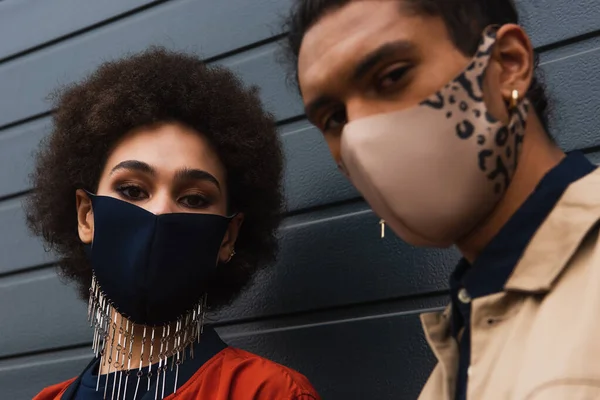 Ricci donna africana americana e uomo sfocato in eleganti maschere protettive in posa al di fuori — Foto stock
