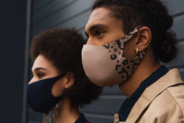 Mujer afroamericana borrosa y hombre joven con elegantes máscaras protectoras afuera - foto de stock