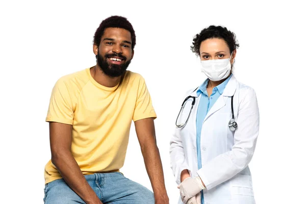 Sorridente afroamericano paziente e medico in maschera medica guardando la fotocamera isolata su bianco — Foto stock