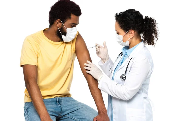 Африканский американец в медицинской маске сидит рядом с врачом со шприцем во время вакцинации изолирован на белом — стоковое фото