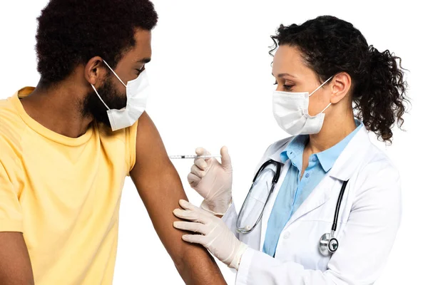 Doente afro-americano sentado ao lado do médico com máscara médica segurando seringa durante a vacinação isolada em branco — Fotografia de Stock