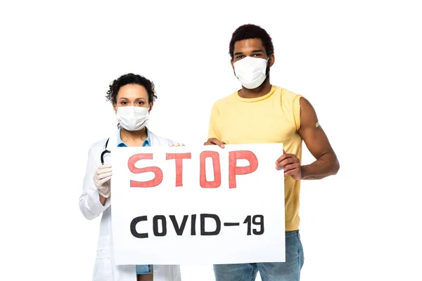 Paciente afroamericano y médico sosteniendo pancarta con letras stop covid-19 aisladas en blanco - foto de stock