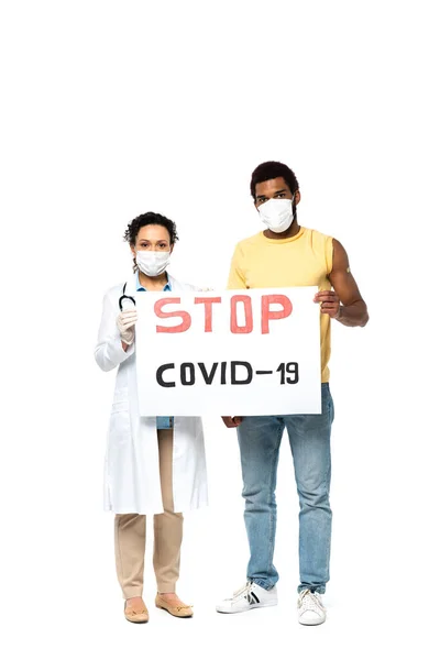 Médico afroamericano y paciente con máscaras médicas sosteniendo pancarta con letras stop covid-2019 sobre fondo blanco - foto de stock