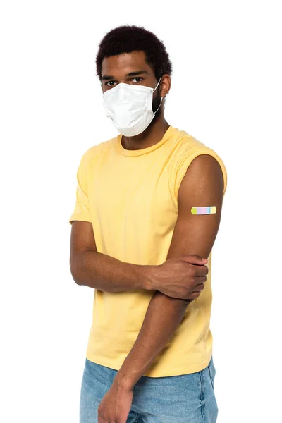 Afro-Américain en masque médical et plâtre adhésif sur le bras en regardant la caméra isolée sur blanc — Photo de stock