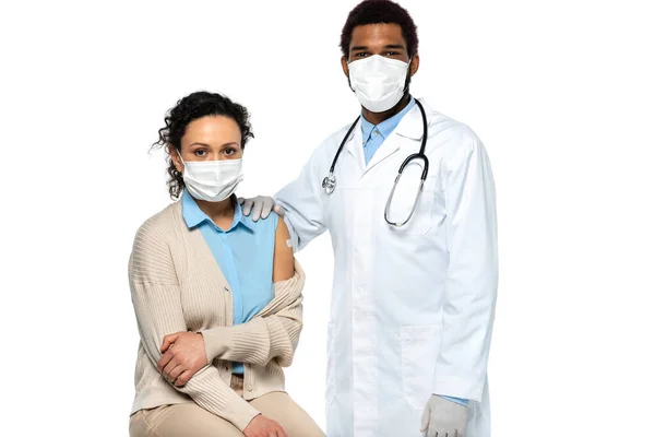 Médico afroamericano en guantes de látex de pie cerca del paciente en máscara médica y parche adhesivo en el brazo aislado en blanco - foto de stock