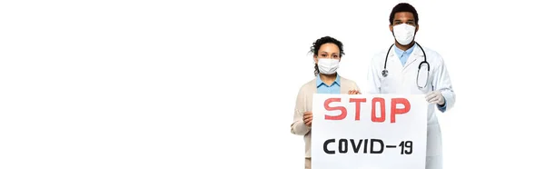 Paciente afroamericano y médico en máscaras médicas con pancarta con letras stop covid-2019 aisladas en blanco, pancarta - foto de stock