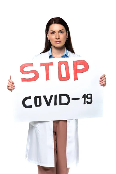 Médico morena mirando a la cámara mientras sostiene la pancarta con stop covid-2019 letras aisladas en blanco - foto de stock