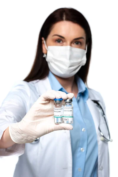 Médico borroso con máscara médica sosteniendo frascos con vacuna aislada en blanco - foto de stock