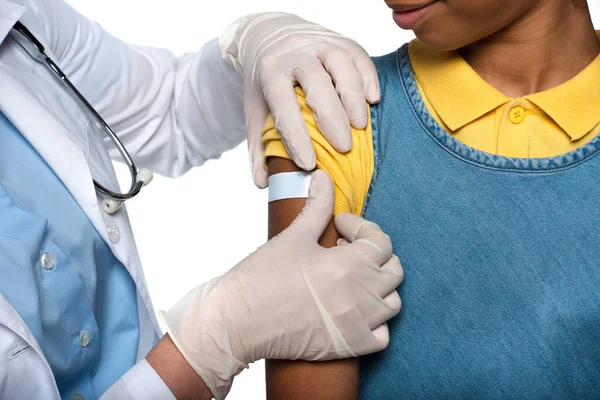 Vista recortada del pediatra que aplica yeso adhesivo en el brazo del paciente afroamericano aislado en blanco - foto de stock