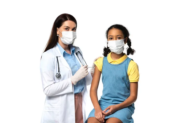 Médico sosteniendo jeringa con vacuna cerca de niño afroamericano en máscara médica aislado en blanco - foto de stock