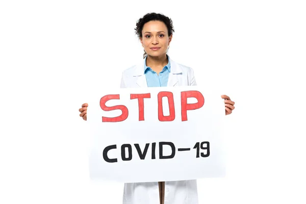 Médico afroamericano sosteniendo pancarta con letras stop covid-2019 y mirando a la cámara aislada en blanco - foto de stock