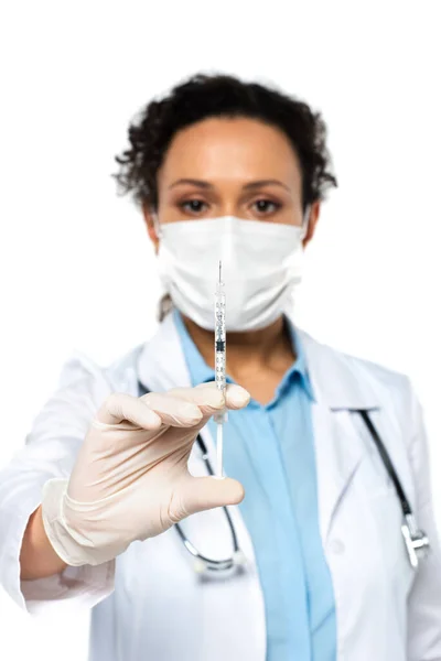 Syringe in hand of african american doctor in medical mask on blurred background isolated on white — Stock Photo
