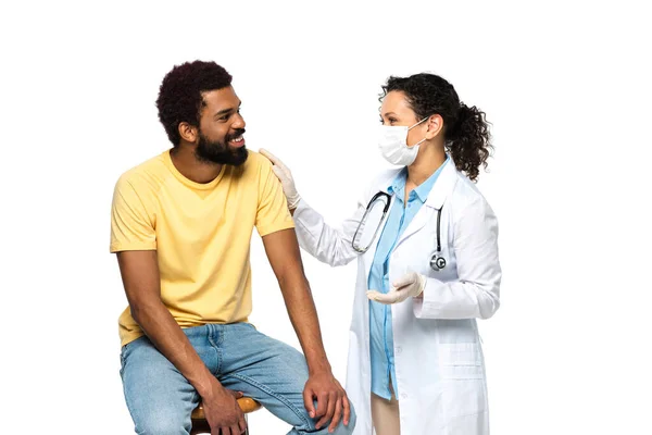 Médico en máscara médica y guantes de látex abrazando paciente afroamericano aislado en blanco - foto de stock